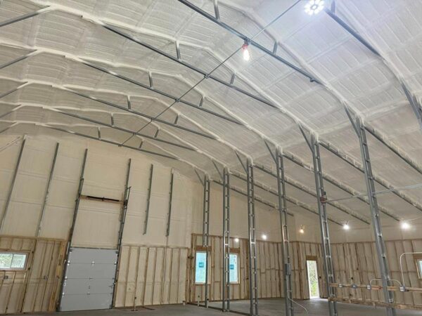 Interior of a large arched metal barn in Onalaska, TX, with freshly applied closed-cell spray foam insulation on the metal roof deck and upper walls.