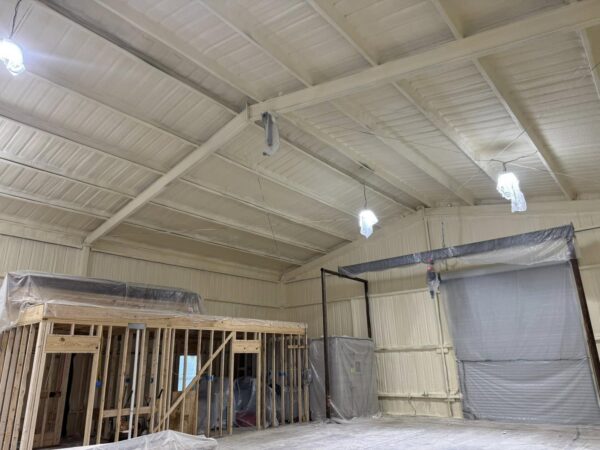 Metal building interior in Livingston, Texas during framing stage with closed cell spray foam insulation applied to corrugated steel walls and roof, wood stud framing and a covered roll-up door visible