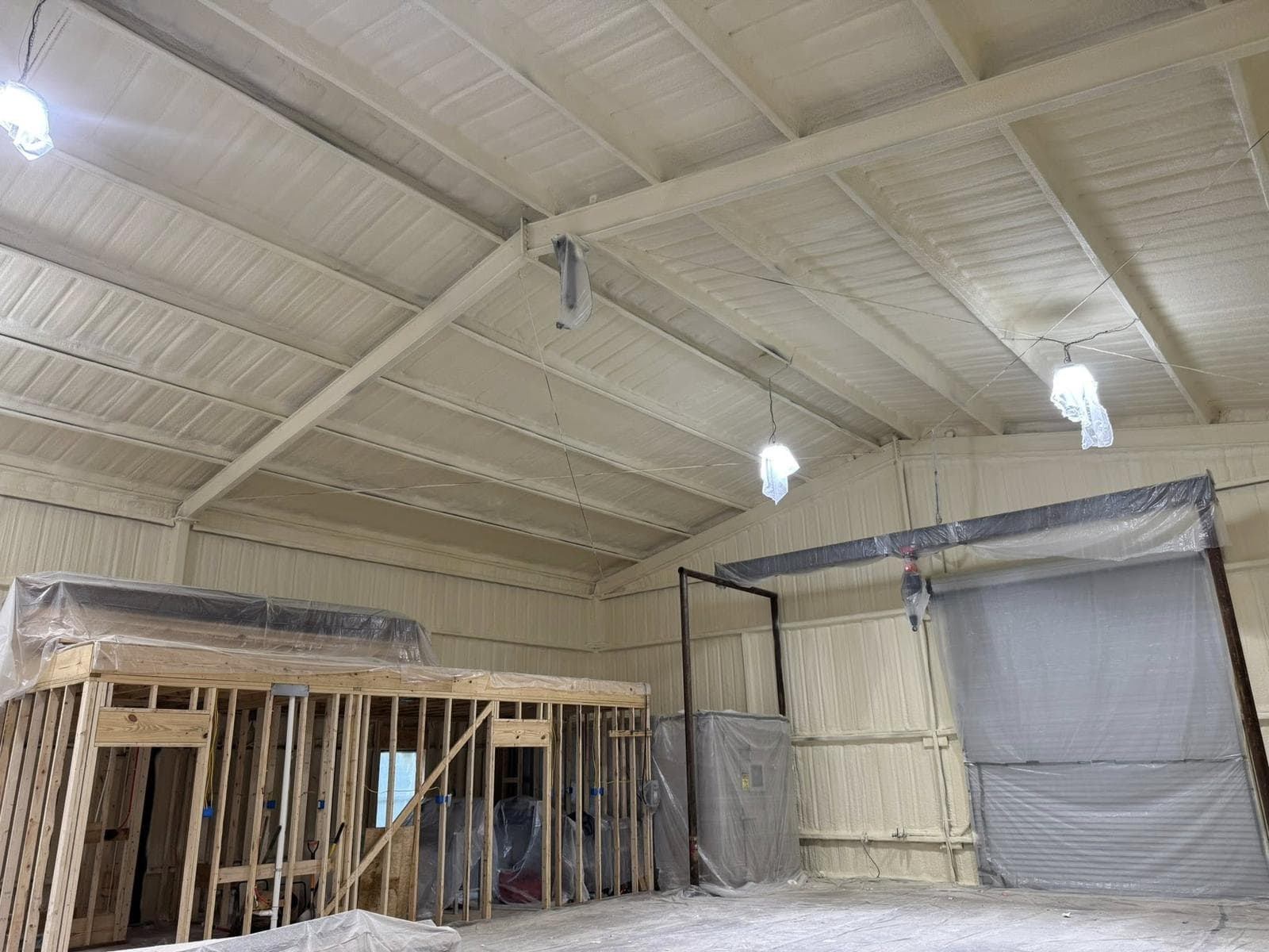 Metal building interior in Livingston, Texas during framing stage with closed cell spray foam insulation applied to corrugated steel walls and roof, wood stud framing and a covered roll-up door visible